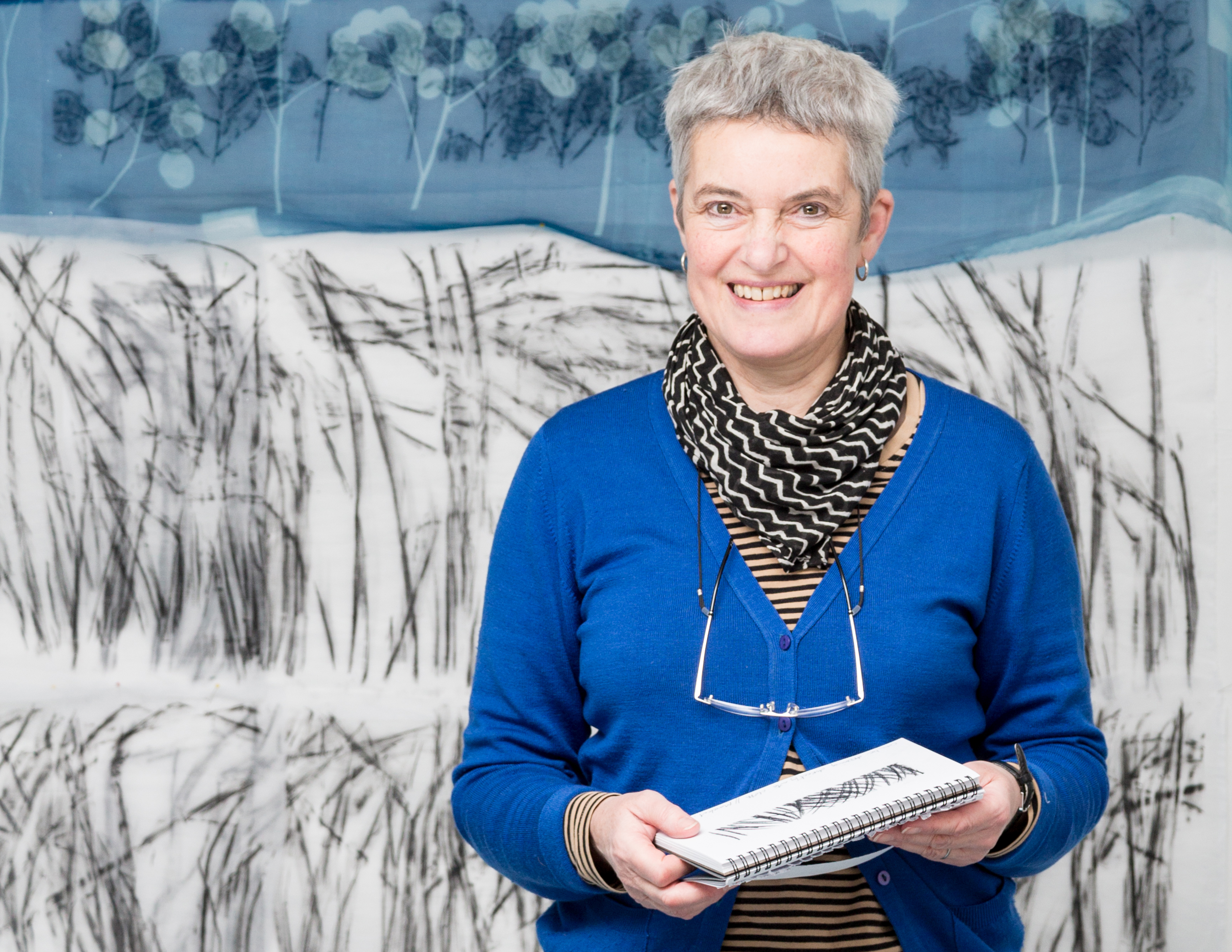 Textile artist and designer Pauline Burbidge stands in front of a quilt. She is wearing a black-and-white striped shirt, blue sweater and black-and-white scarf, with glasses hanging from her neck, holding a sketchpad.