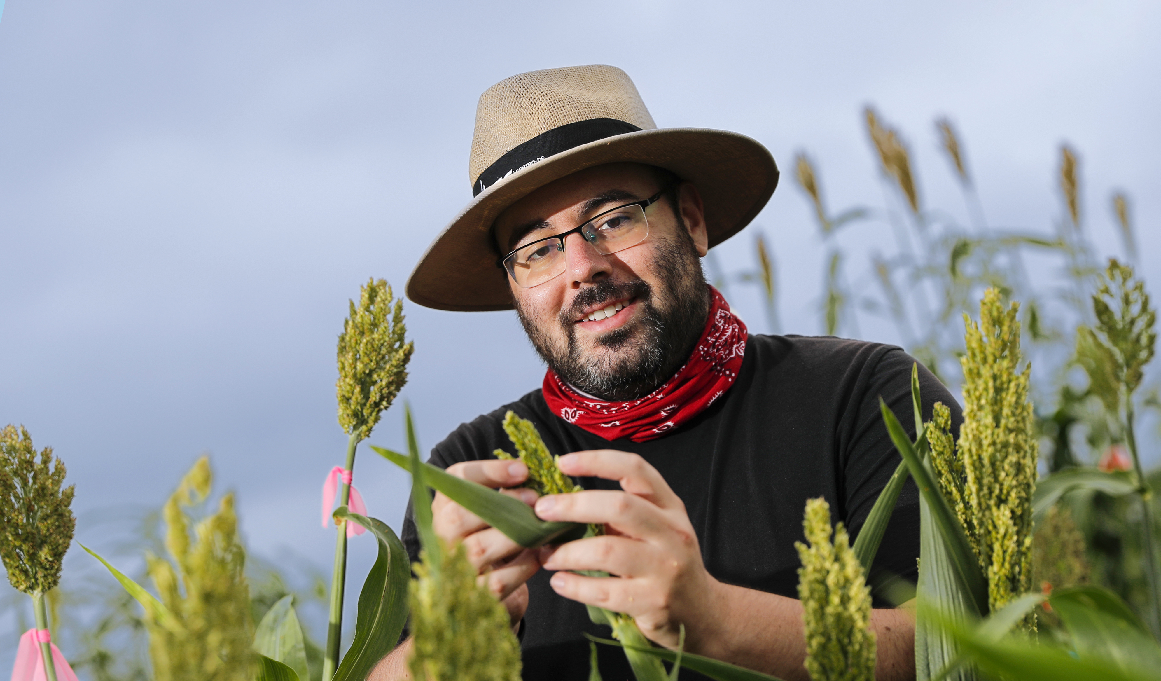 James Schnable stands in a sorghum field. He is wearing a black shirt, red gaiter, wide-brimmed hat and glasses.