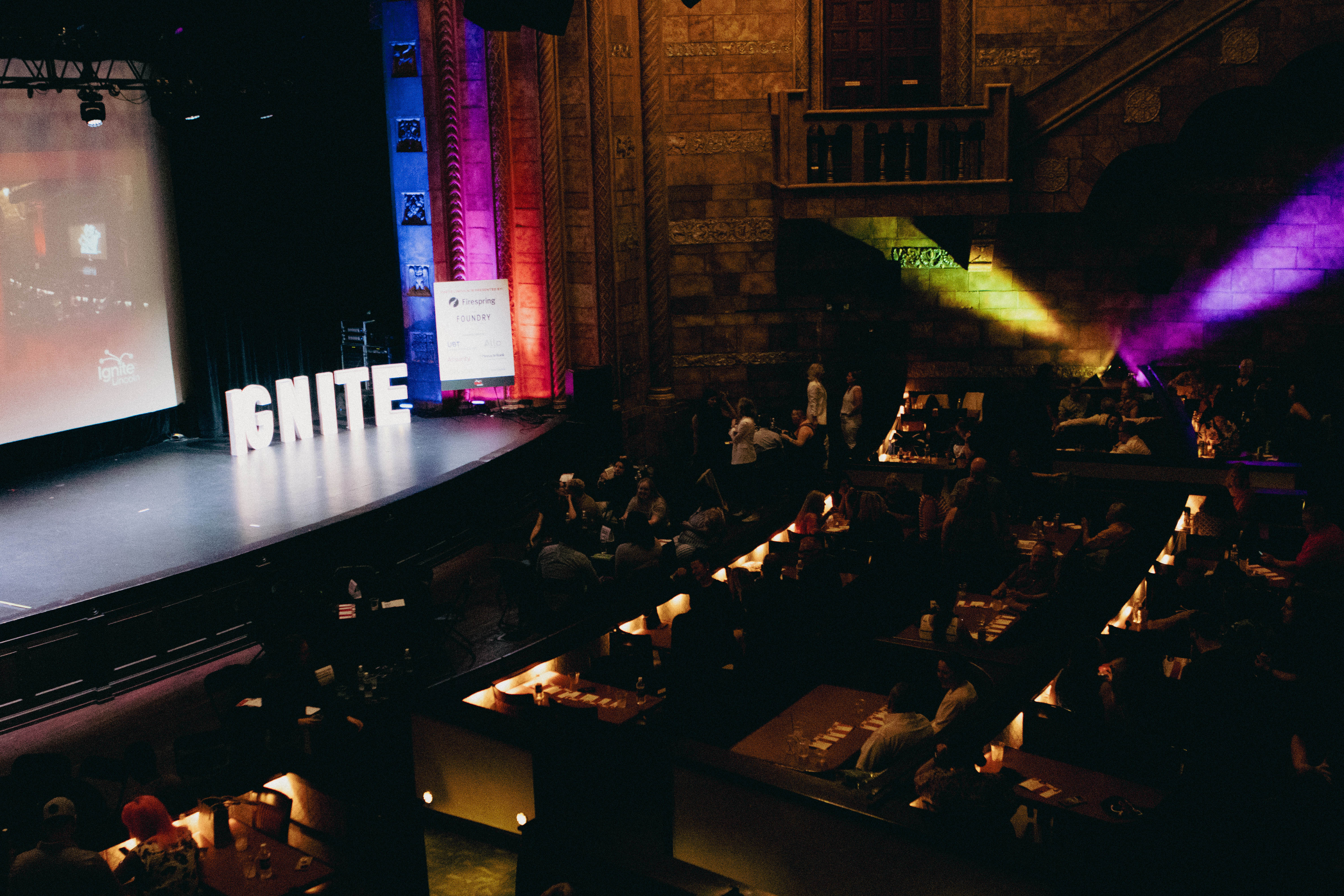 The word "Ignite" graces an empty stage as people wait at tables in the Rococo Theatre.