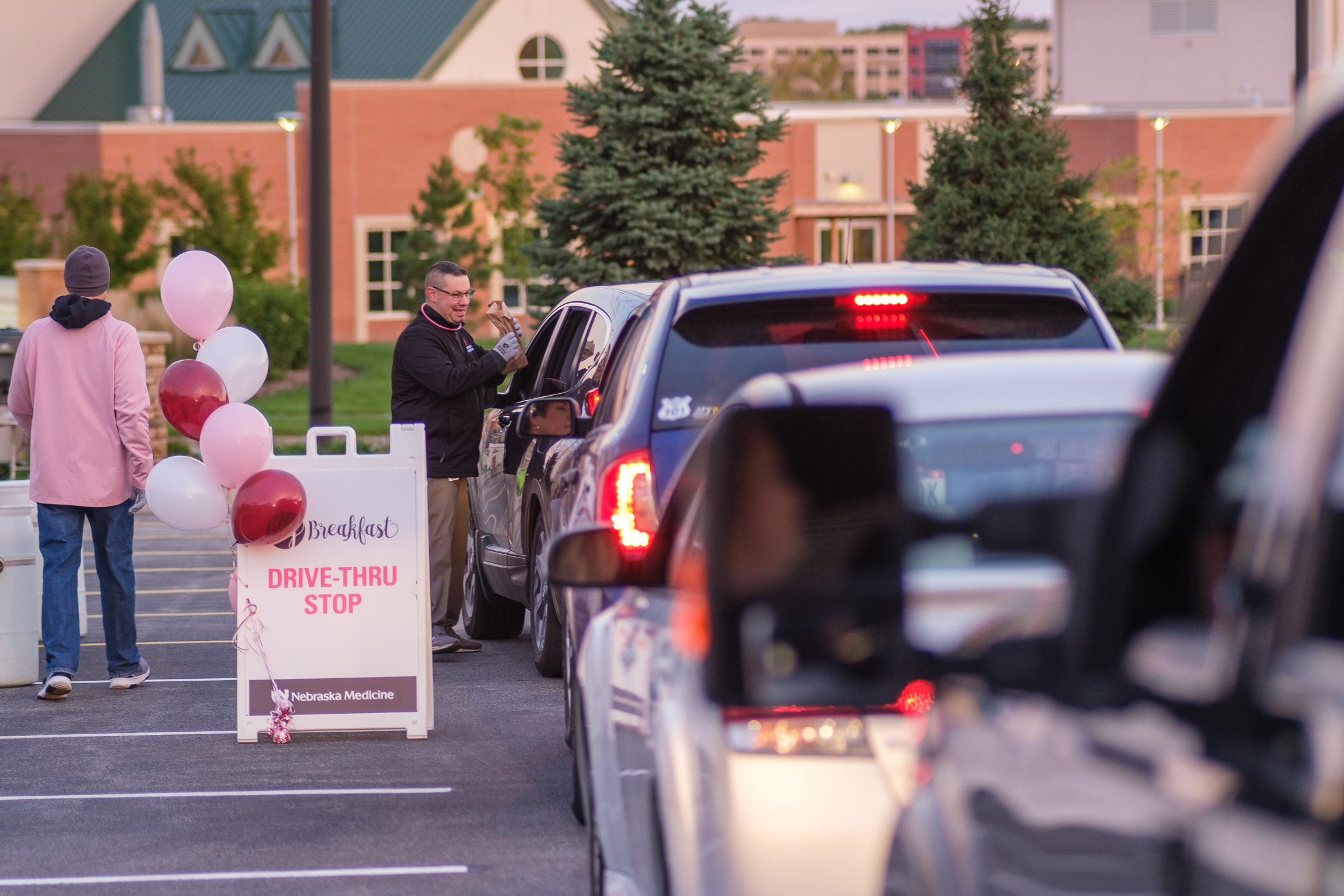 A man hands a breakfast bag through a car window in a parking lot. Several other vehicles are lined up behind the car.