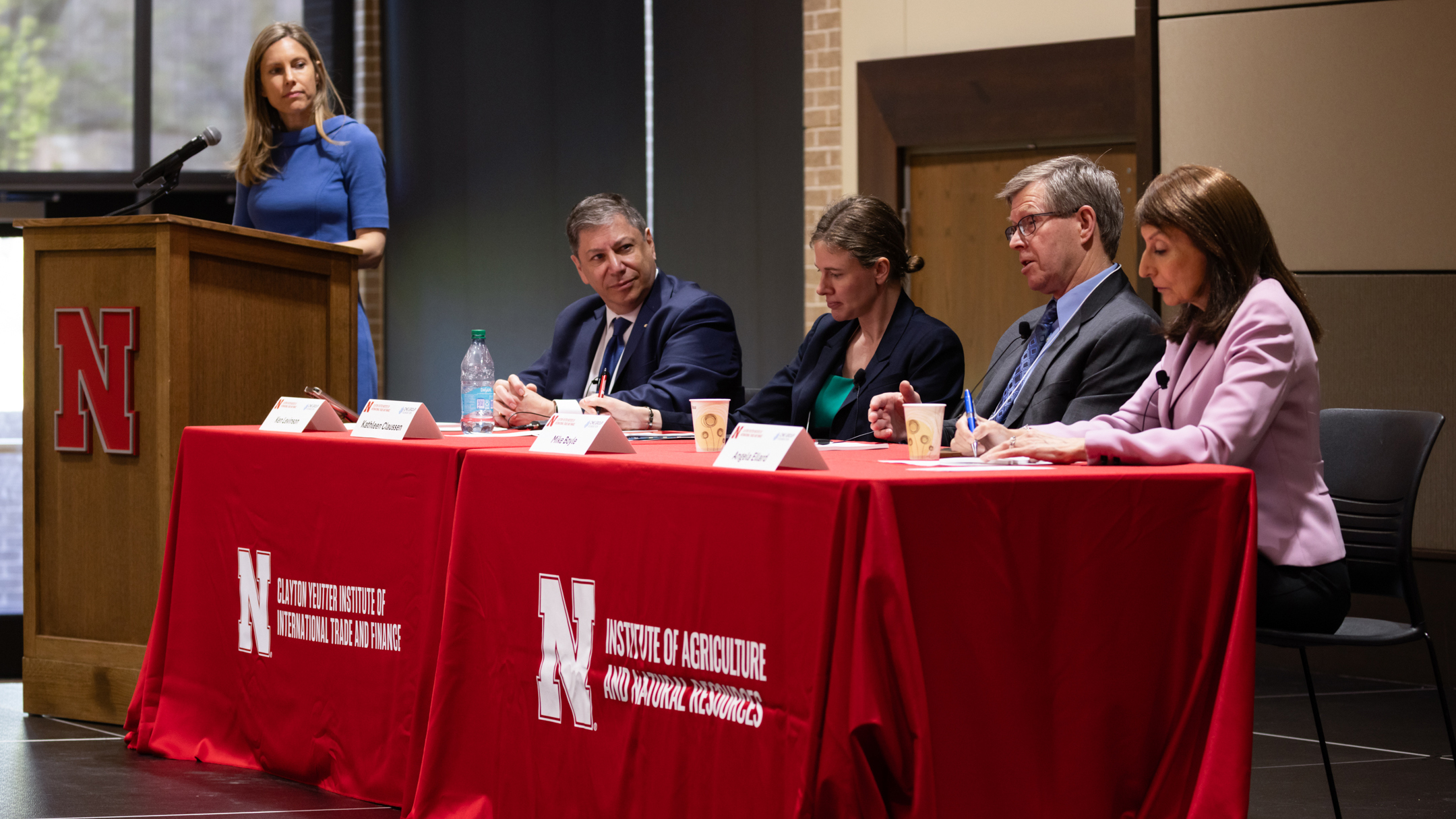 A woman stands at a lectern next to two red cloth-draped tables behind which two men and two women are seated. The table coverings read "Clayton Yeutter Institute of International Trade and Finance" and "Institute of Agriculture and Natural Resources," with white Nebraska N's next to the words.