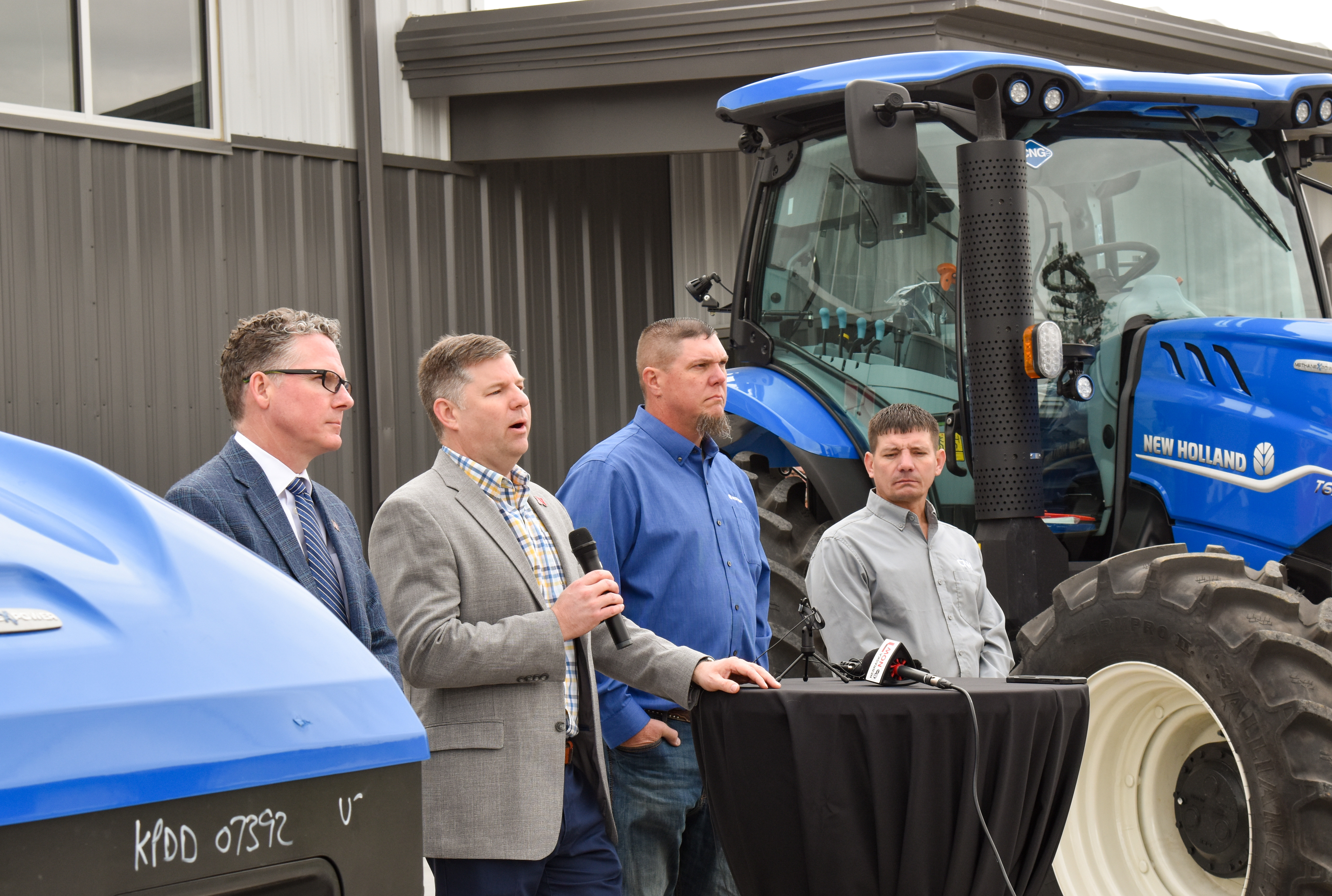A man speaks into a microphone as he stands next to three other men between two New Holland methane-powered tractors.