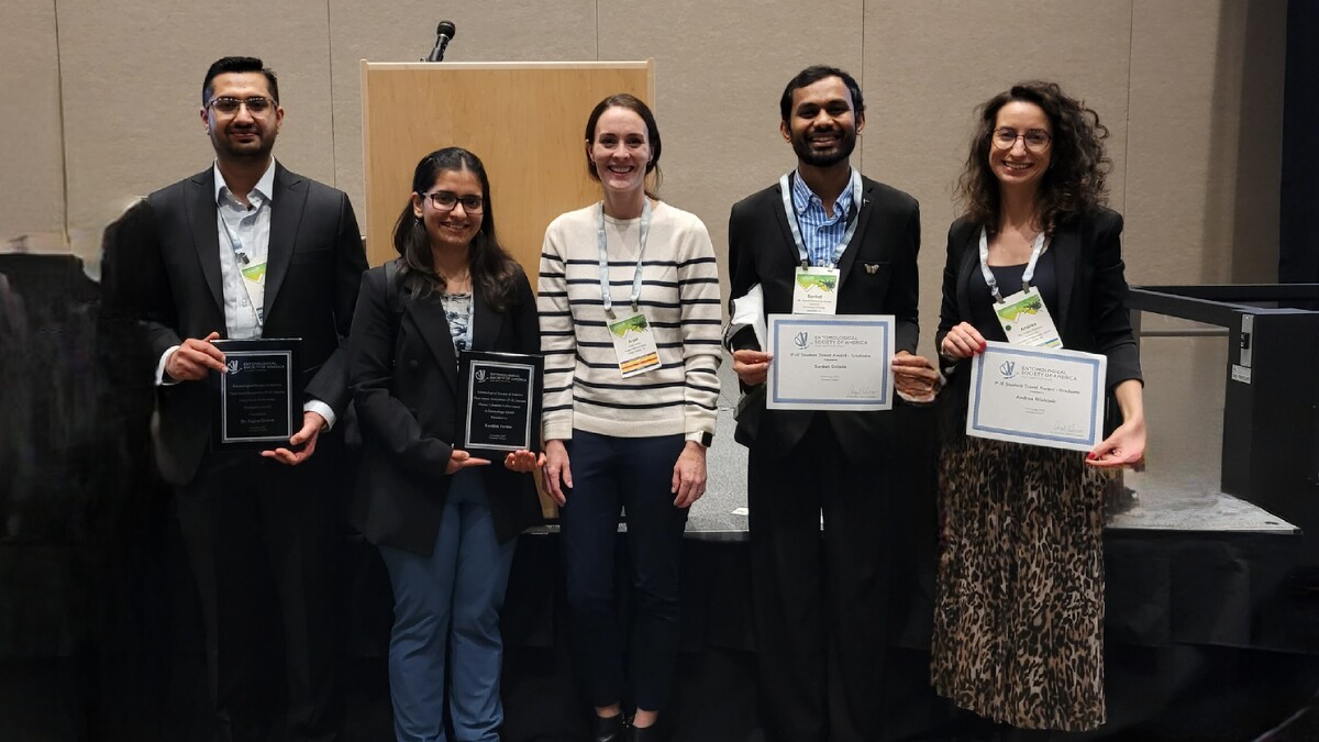 Five award recipients and a presenter pose on a stage at a national entomology conference. From left to right: Sajjan Grover and Kashish Verma hold plaques, Anjel Helms stands in the center as the awards presenter, and Sanket Shinde and Andrea Rilakovic hold certificates.
