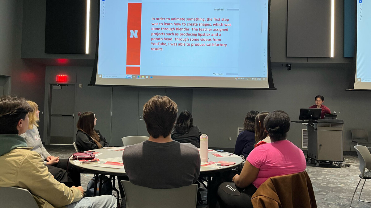 Handsone Tong, a chemistry major and First Year Research Experience student, presents at a lectern as eight other FYRE students gathered at a round table list.