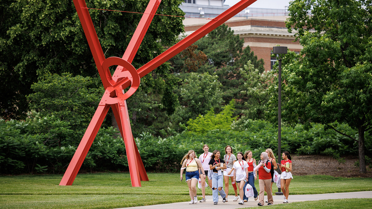 Students walk along a campus path near a red Old Glory sculpture, surrounded by green grass and trees.