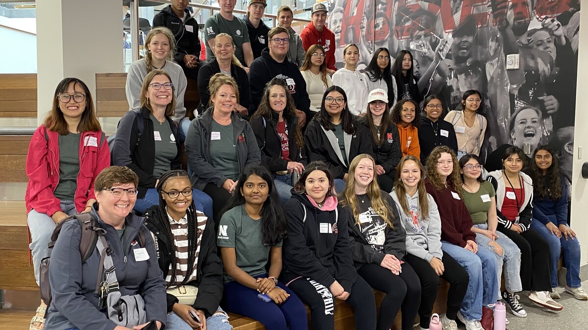 Group photo of Cultivate ACCESS participants, including high school scholars, teachers, UNL mentors, and program staff, seated on indoor steps at the East Campus Union. Everyone is smiling and facing the camera. A “Go Huskers” mural is visible in the background.