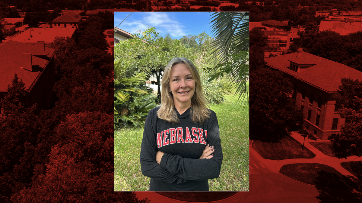 A woman stands outdoors in a lush, green garden with trees and tropical plants behind her, smiling at the camera with her arms crossed. She is wearing a black long-sleeve shirt with “Nebraska” in red lettering. The image is framed over a red-tinted aerial view of the University of Nebraska–Lincoln campus.