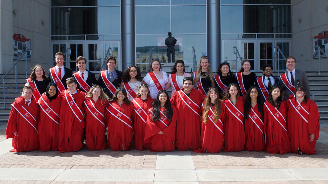 Thirteen University of Nebraska–Lincoln students — all new members of the Innocents Society — wear red robes with red-and-white sashes in the front row of a group photo on the east side of Memorial Stadium. In the back row are 12 other Huskers students — all 2024-25 members of the Innocents Society — wearing formal attire with red-and-white sashes.
