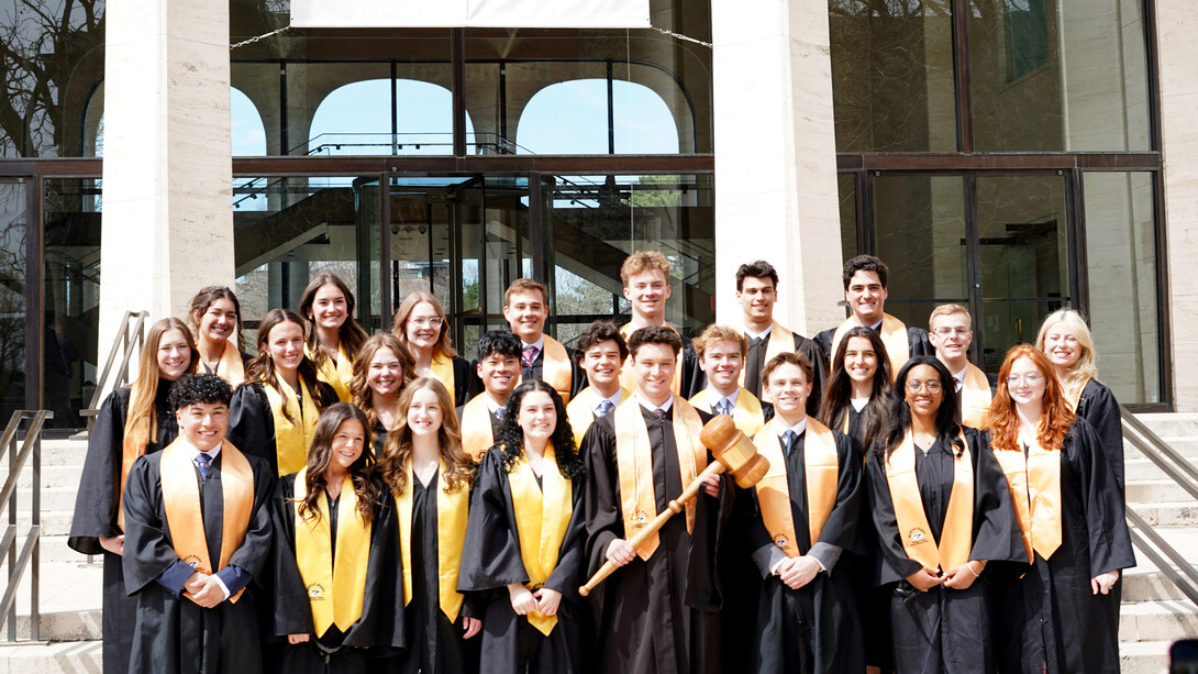 Twenty-four University of Nebraska–Lincoln students — all new members of the Black Masque Chapter of Mortar Board — pose together in black robes with gold stoles on the front steps of Sheldon Museum of Art. A young man in the front row holds an oversized gavel.