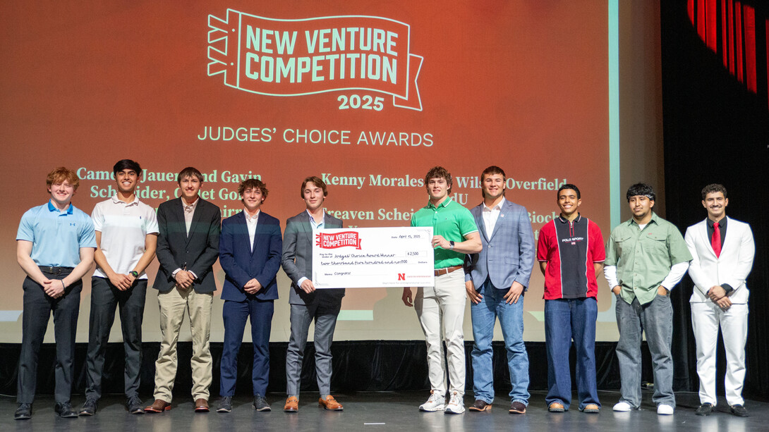 Ten young men stand in a row on stage. The two in the middle hold an oversized check. They stand in front of a screen that reads "New Venture Competition 2025: Judges' Choice Awards."