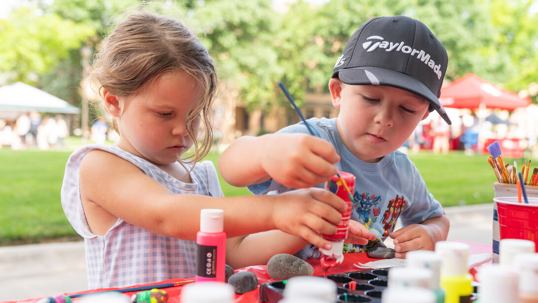 A young girl and young boy paint rocks at an outdoor festival table.