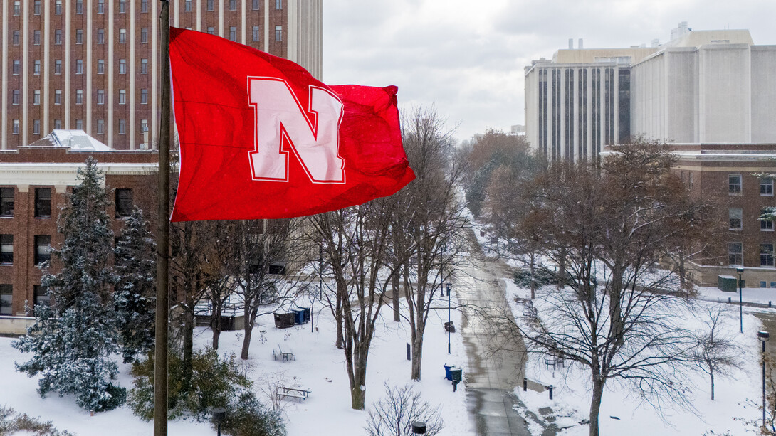 A red flag with a white Husker N waves over a sidewalk on the University of Nebraska–Lincoln's City Campus. Snow covers the ground.