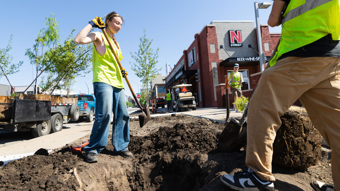 A woman stands next to a hole where a tree will be planted.