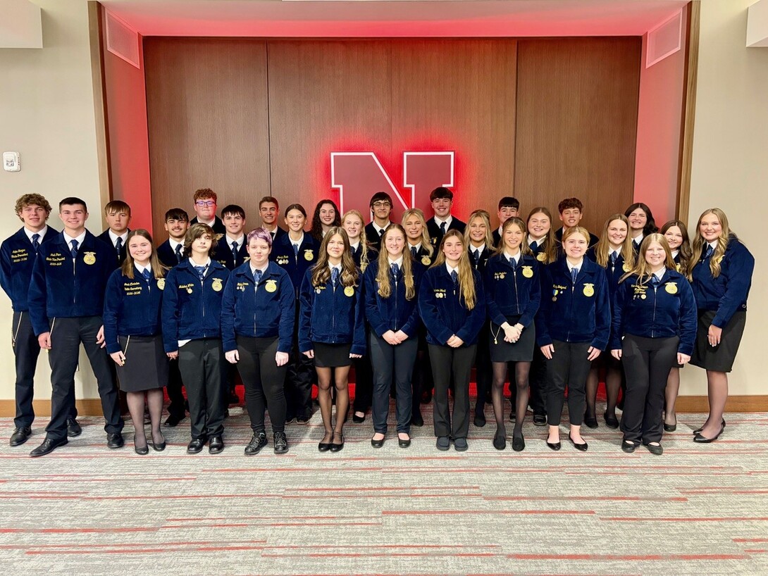 Group of Nebraska FFA students wearing official blue jackets stand together for a photo inside the University of Nebraska–Lincoln, with a large illuminated red “N” behind them.