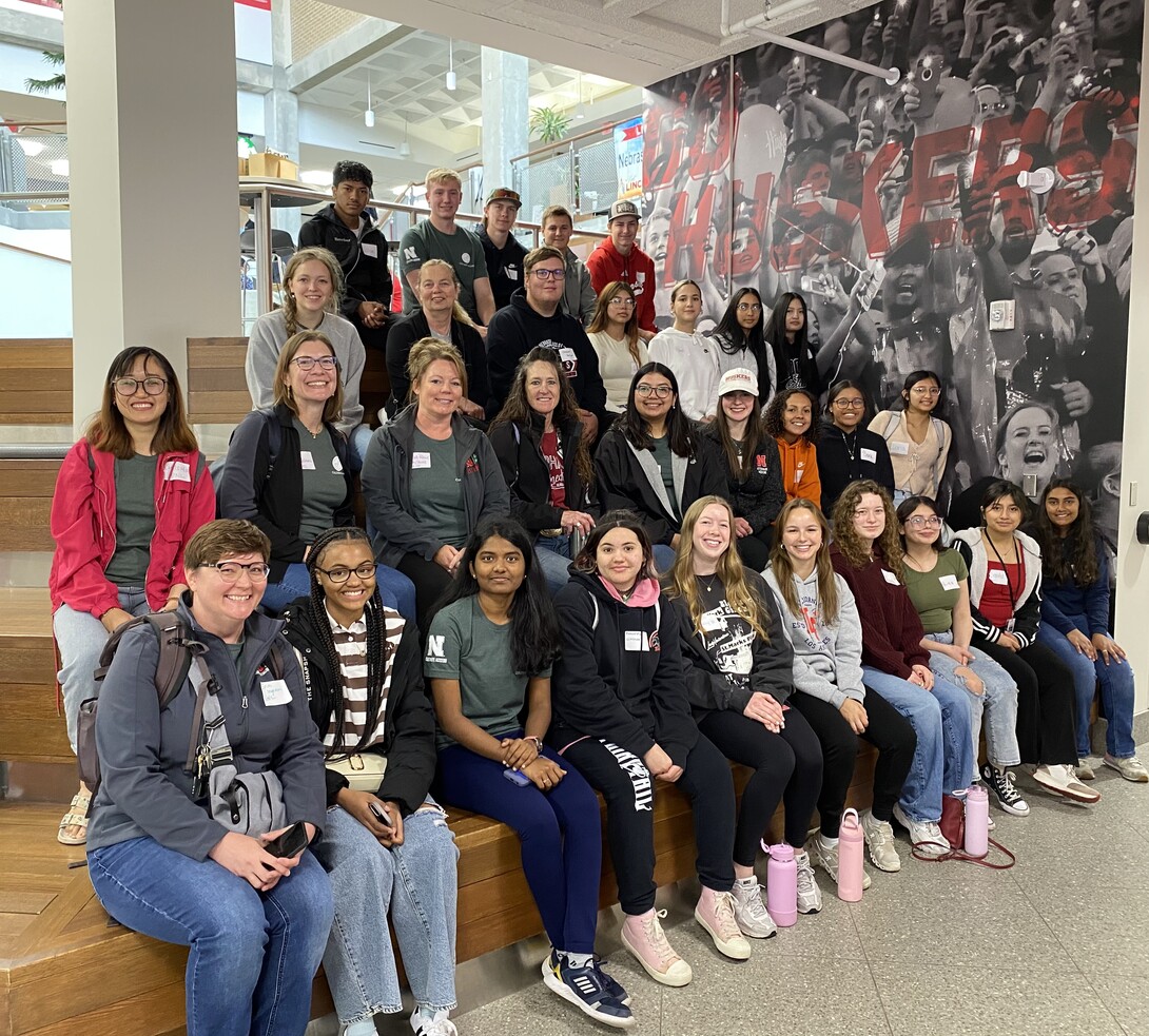Group photo of Cultivate ACCESS participants, including high school scholars, teachers, UNL mentors, and program staff, seated on indoor steps at the East Campus Union. Everyone is smiling and facing the camera. A “Go Huskers” mural is visible in the background.