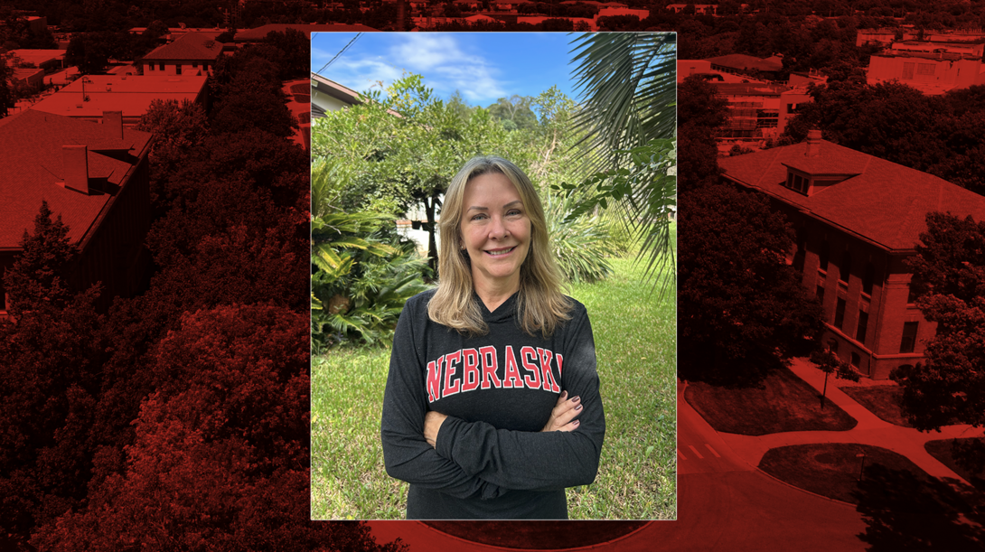 A woman stands outdoors in a lush, green garden with trees and tropical plants behind her, smiling at the camera with her arms crossed. She is wearing a black long-sleeve shirt with “Nebraska” in red lettering. The image is framed over a red-tinted aerial view of the University of Nebraska–Lincoln campus.