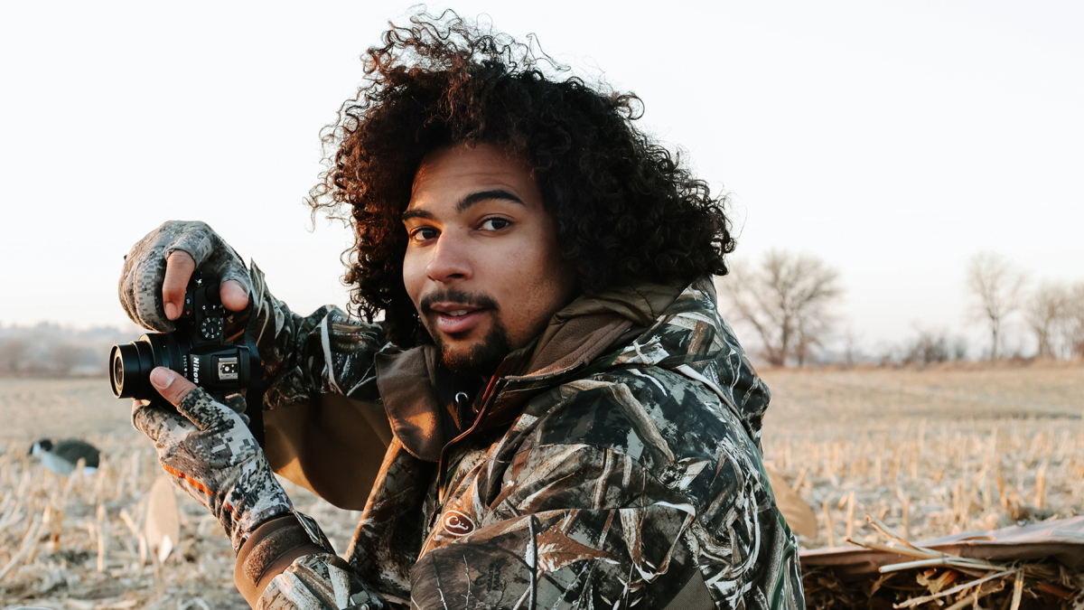 Photo of Elijah Riley in camouflage gear, holding a camera and taking photos outdoors.