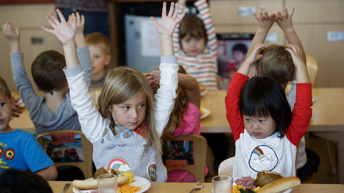 Young children raise their hands in a lunchroom.